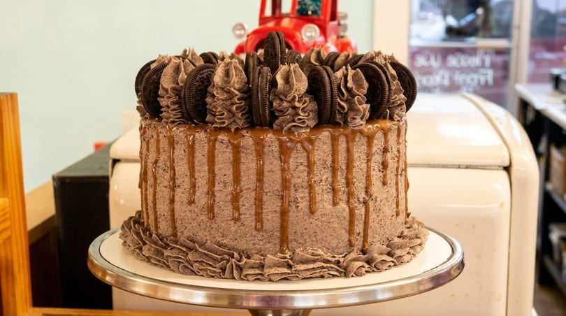 Chocolate cake with caramel drip, topped with Oreo cookies and chocolate frosting swirls on a stand.