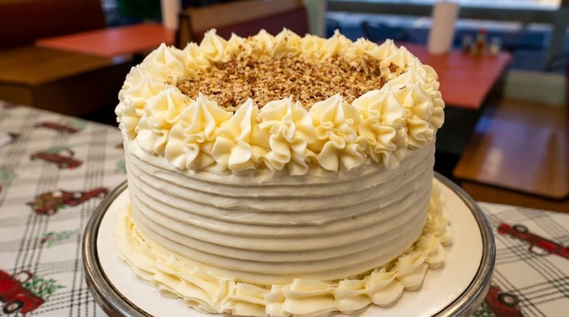 White frosted cake with ridged sides, piped border, and chopped nuts on a festive tablecloth.