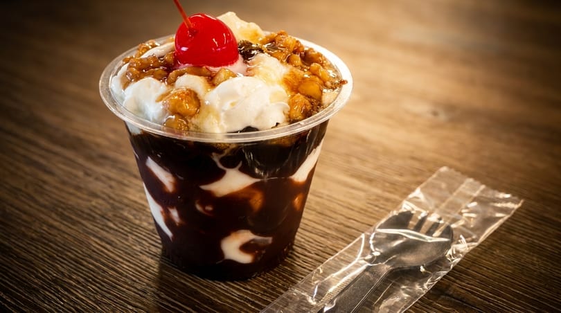 Chocolate sundae with whipped cream, nuts, and a cherry in a cup on a wooden table with wrapped cutlery.