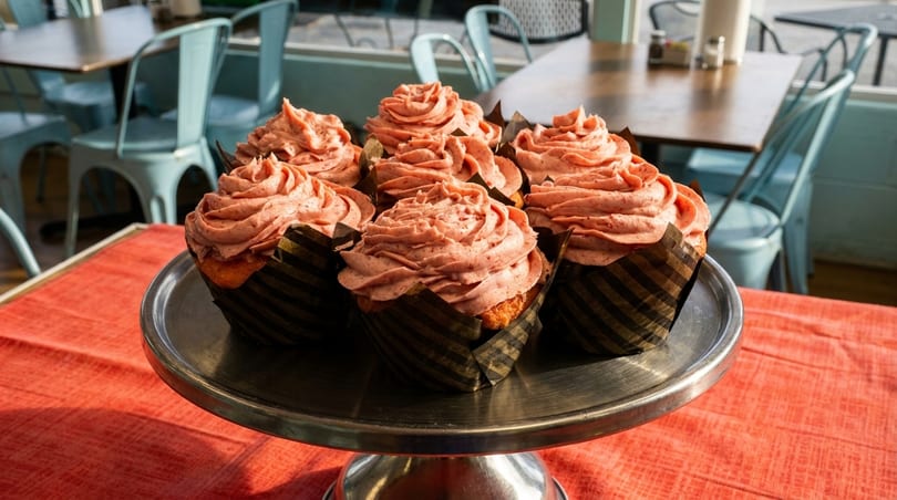 Seven pink-frosted cupcakes on a silver stand in a restaurant with light blue chairs.
